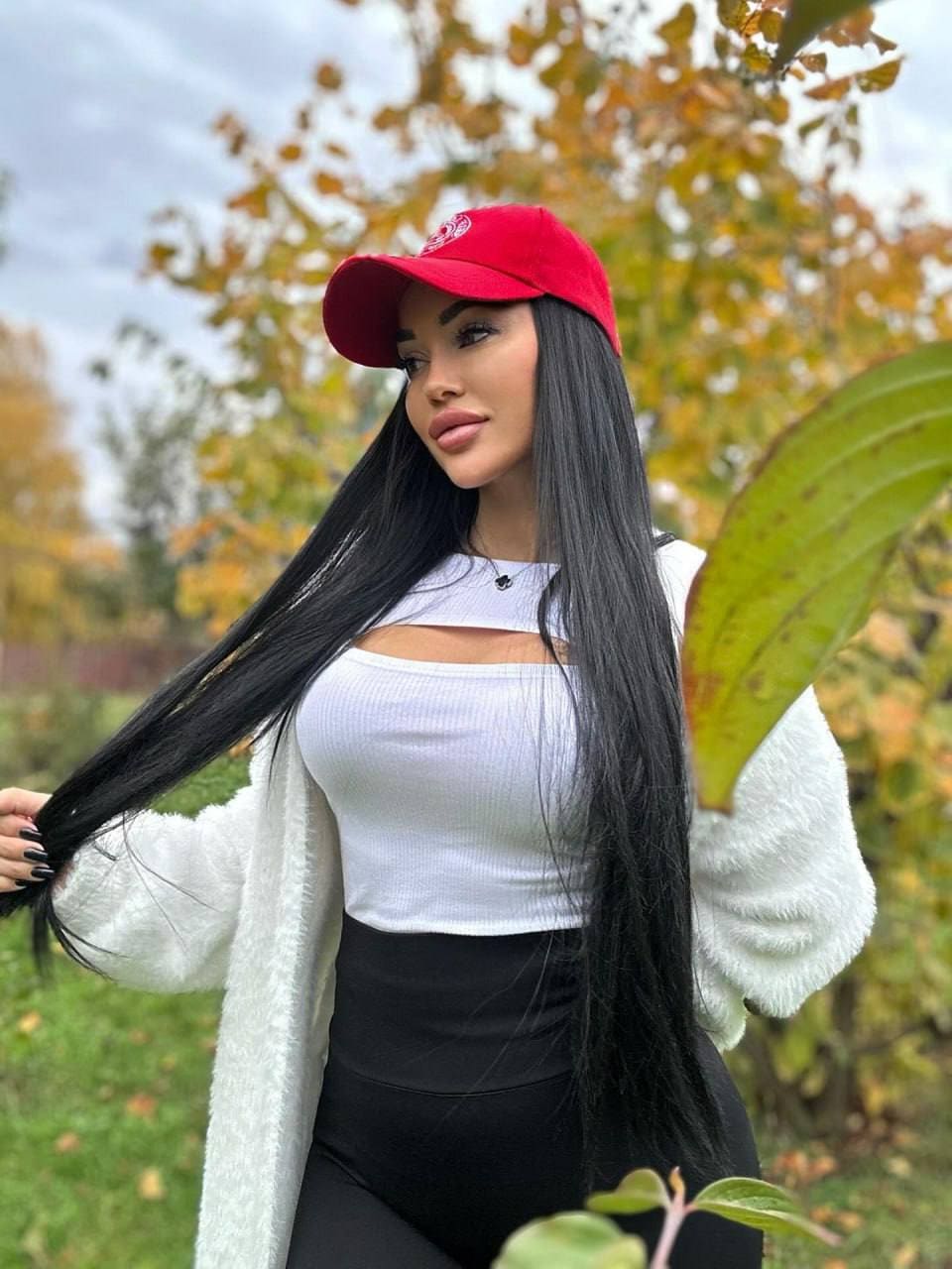 A woman in a red cap, white top, and fuzzy cardigan stands among autumn foliage outdoors, holding a strand of hair.