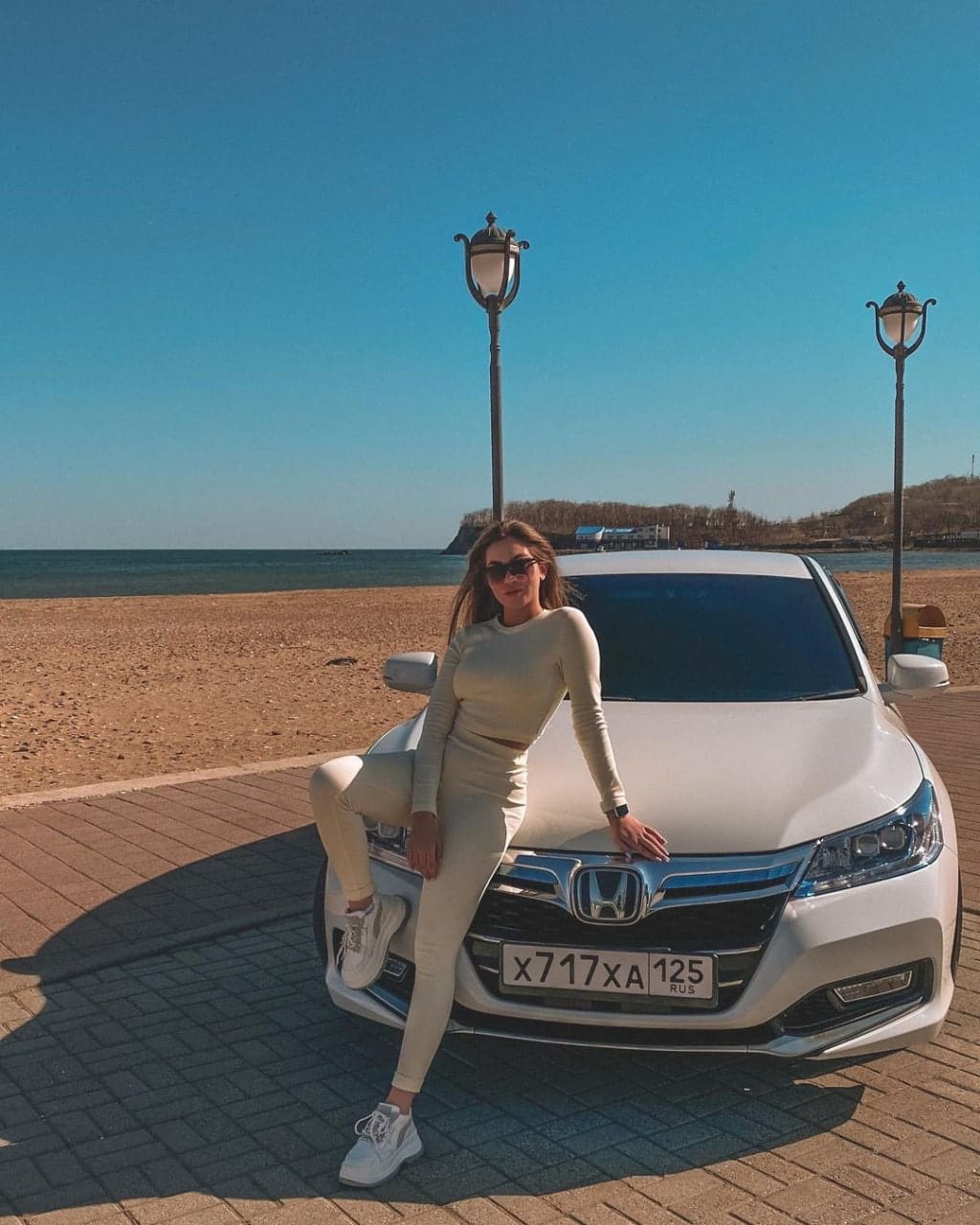 Женщина в очках и кремовом костюме опирается на белый автомобиль на набережной, за ней пляж и море.