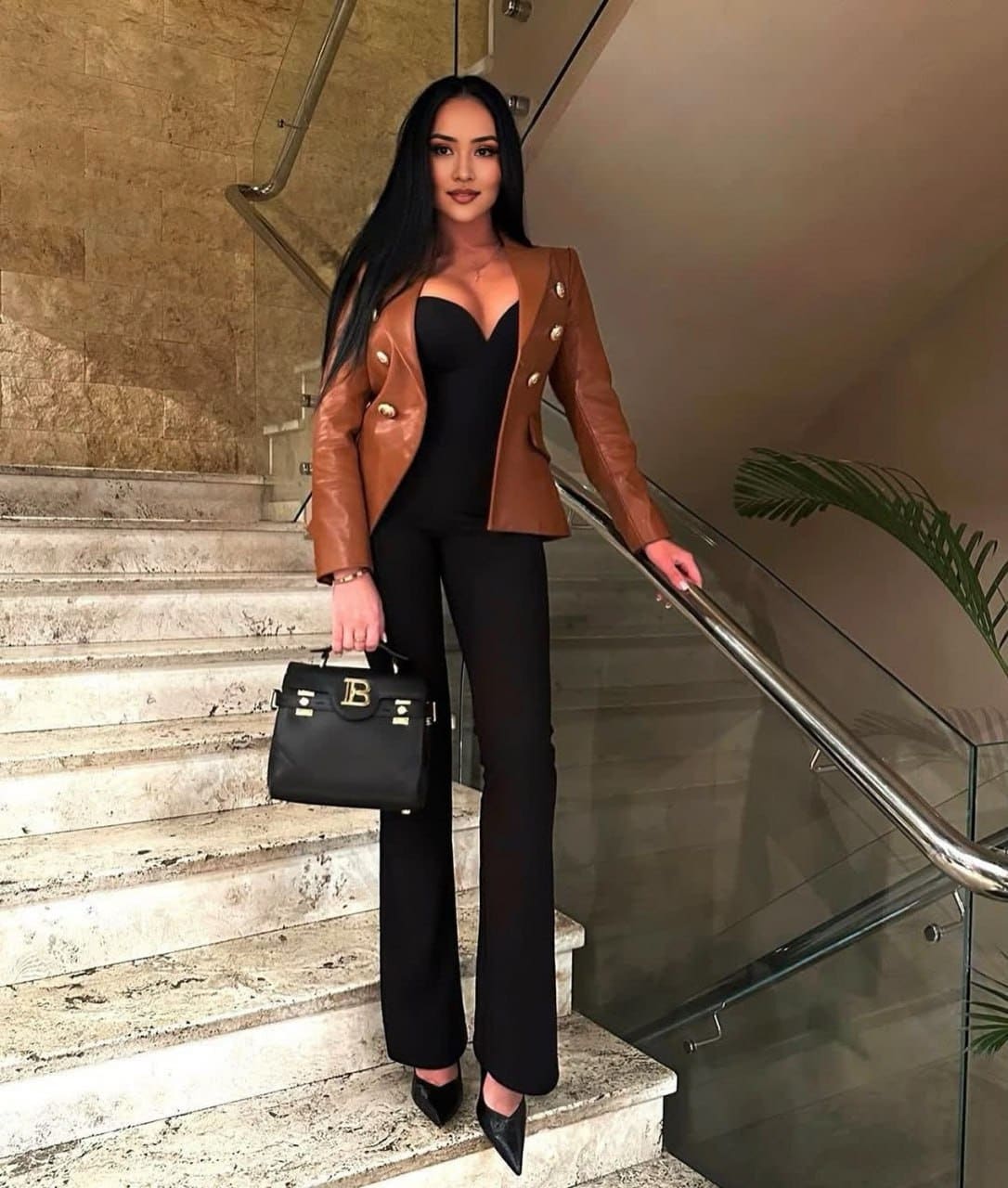 On a marble staircase, a woman in a brown leather jacket, black top, and trousers holds a black handbag while resting a hand on the metal railing.