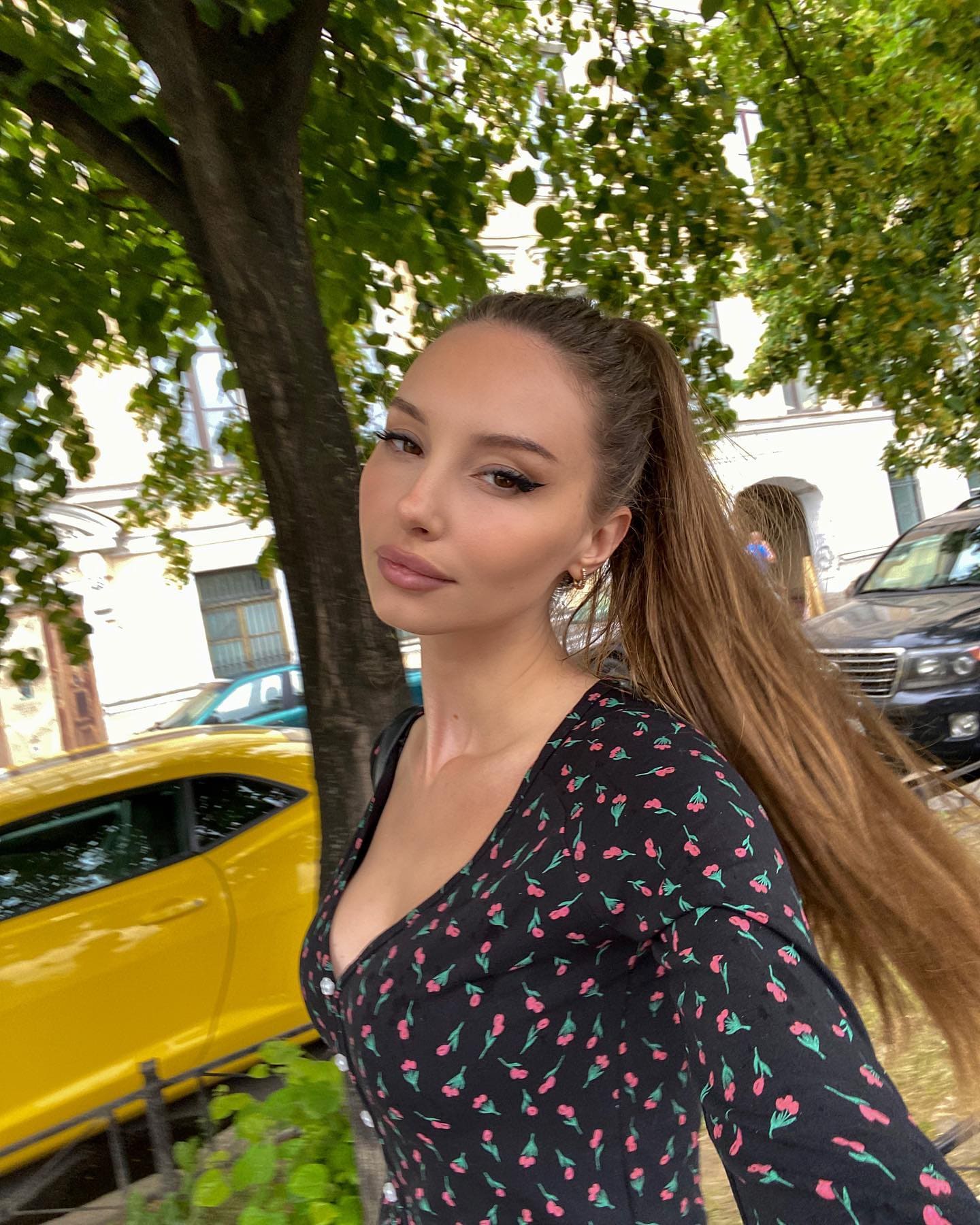 Женщина в черной кофте с принтом вишен и глубоким декольте стоит на улице у дерева, на фоне видны желтый автомобиль и здание.