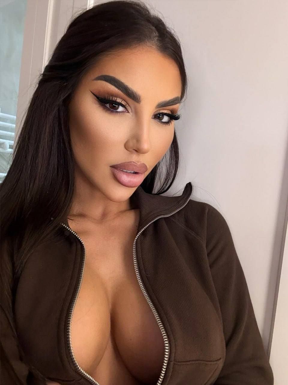 Posing indoors in an unzipped brown jacket against a plain light wall, revealing cleavage with heavy eye makeup.
