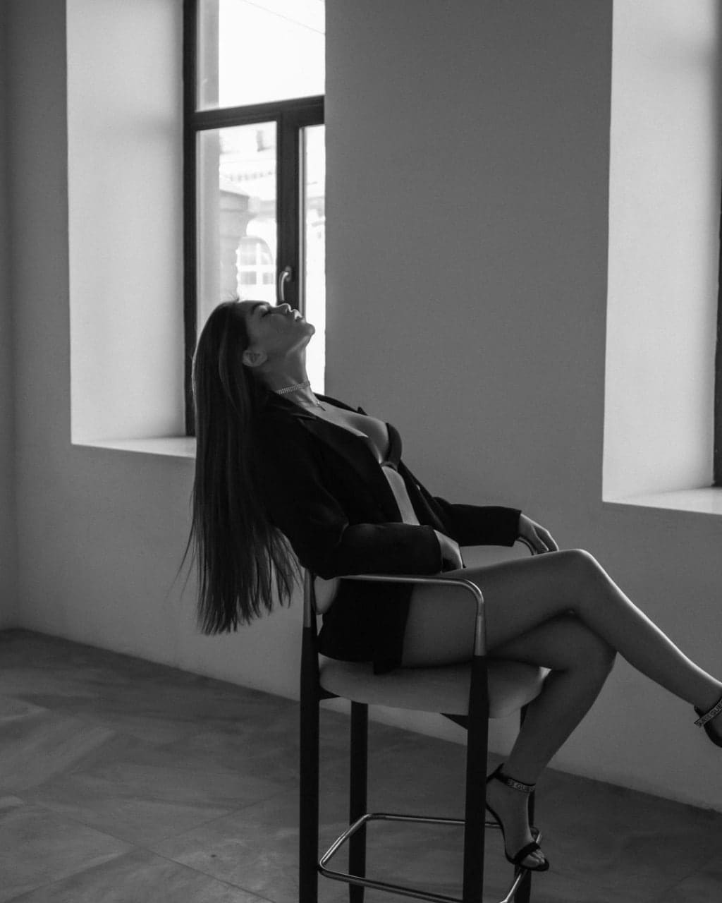 A woman sits on a stool in a black blazer and heels, head tilted back toward a window.