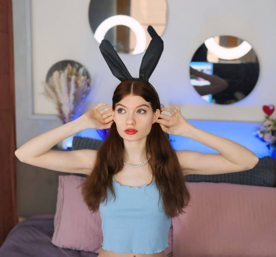 A woman wearing black bunny ears and a light blue cropped tank top poses in a bedroom with round mirrors behind her.