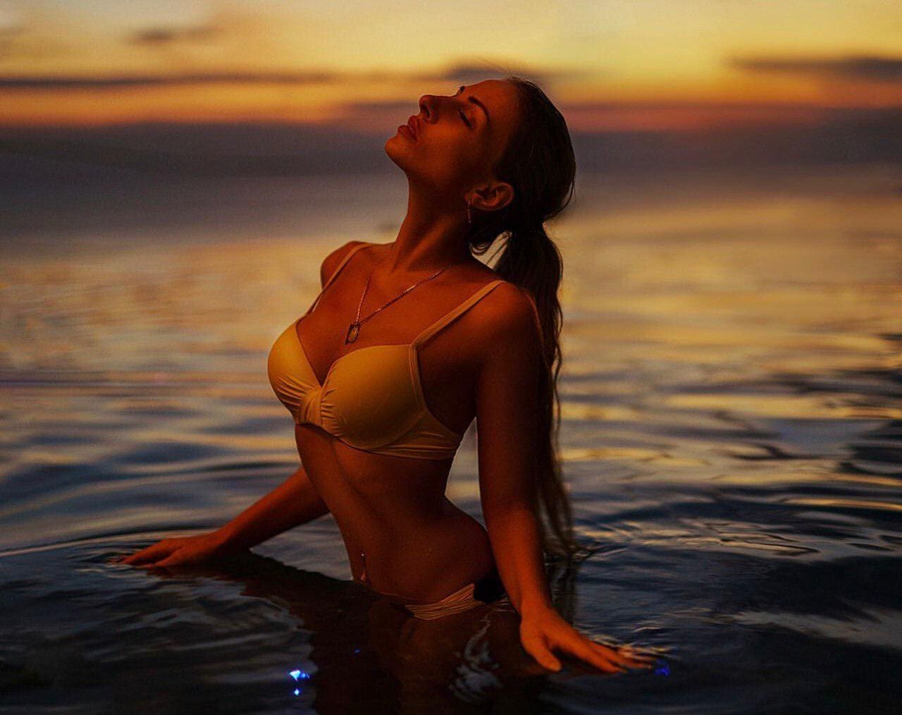 A woman with dark hair tied back sits in water at sunset, wearing a light bikini with her head tilted back and arms resting on the surface.