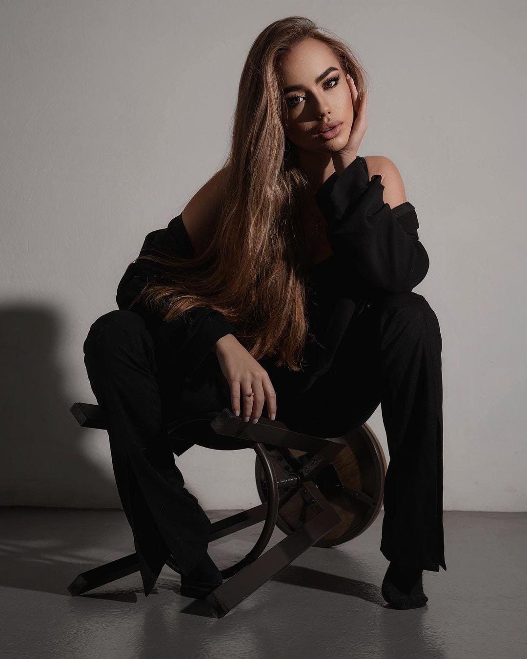 A brunette in a black off-shoulder top sits on a stool, resting a hand on her cheek against a light wall.