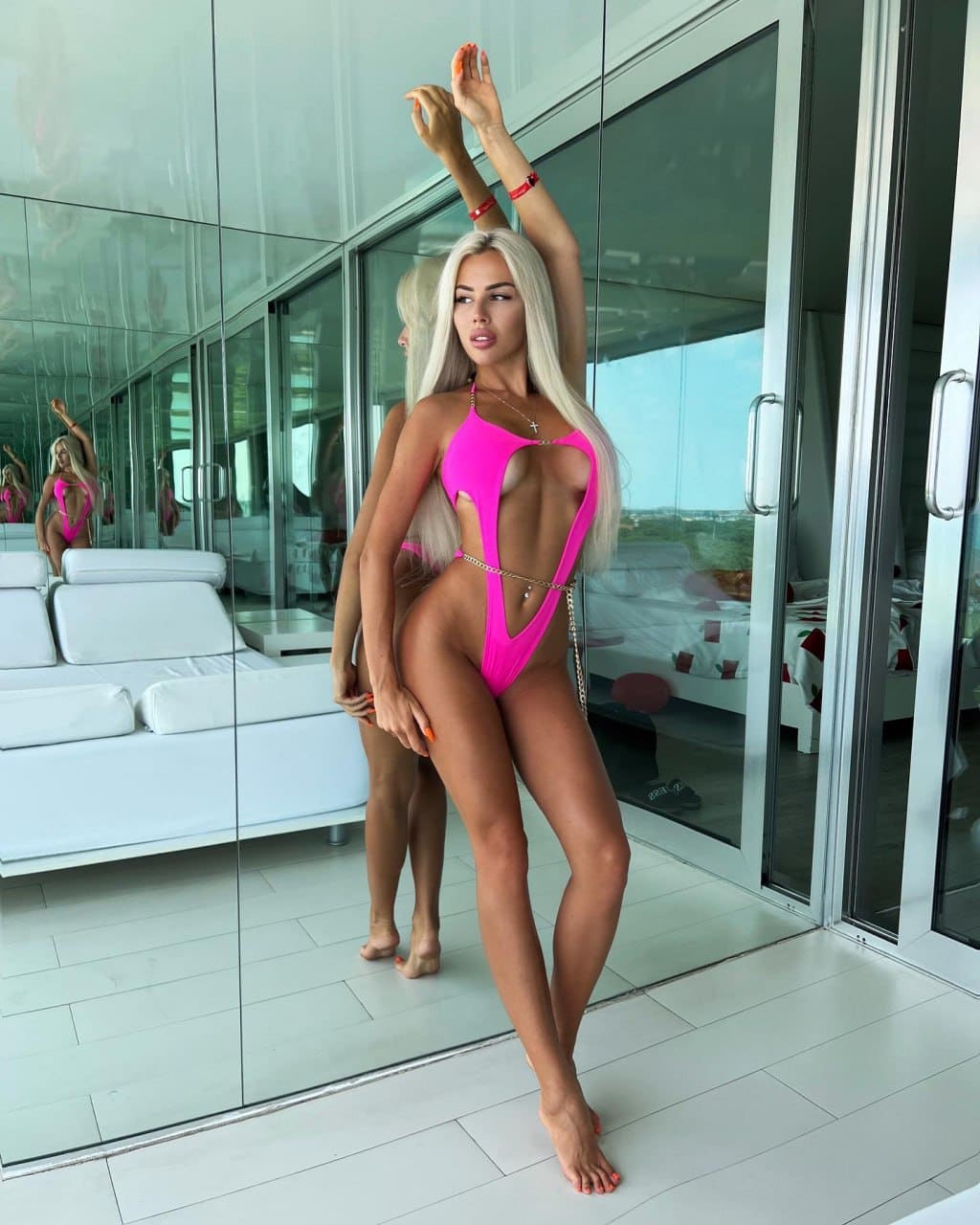 Девушка в розовом слитном купальнике с вырезами и цепочкой стоит у панорамного окна, подняв руку.