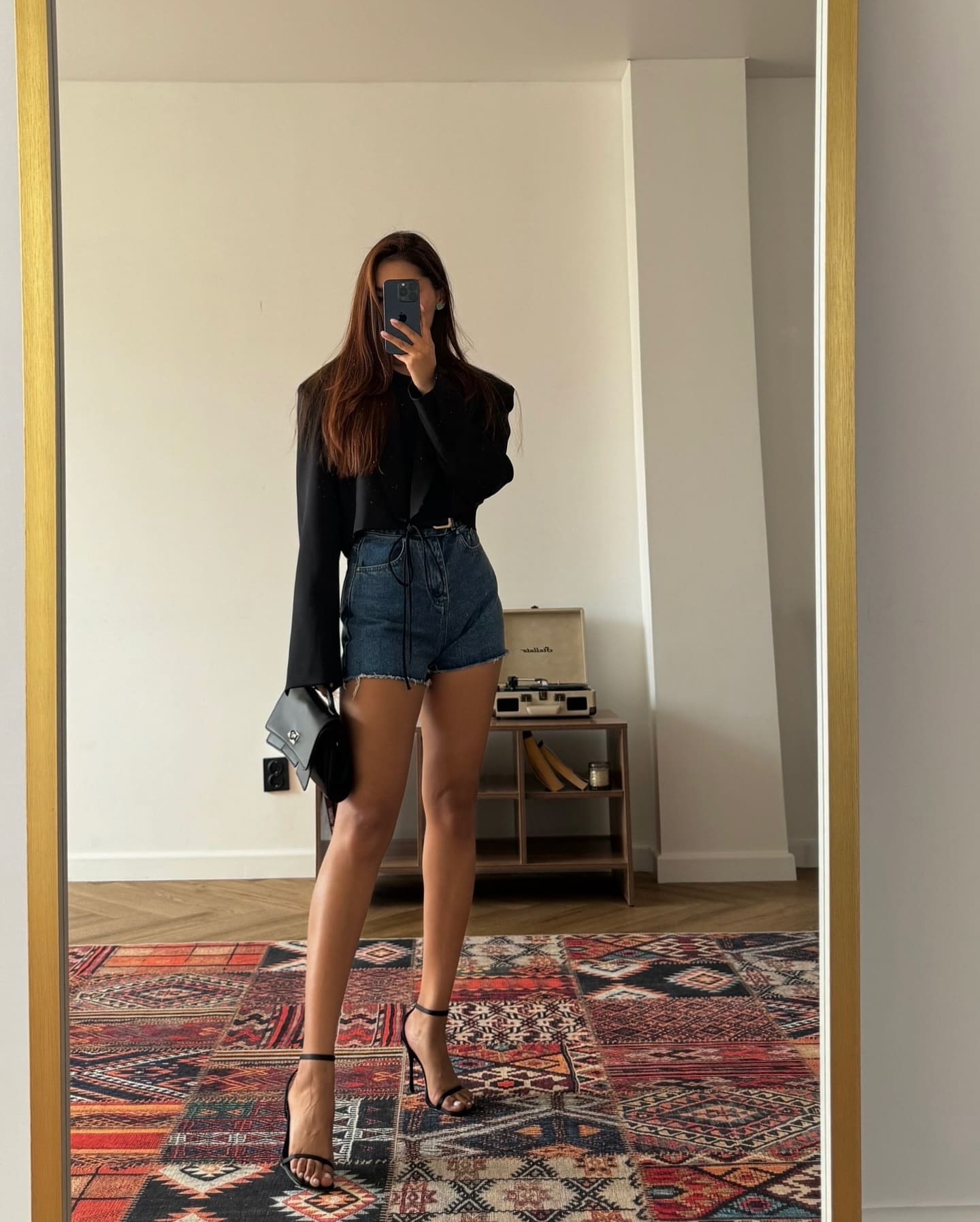 A woman takes a mirror selfie in a black long-sleeved top, denim shorts, and heels, with a patterned rug and record player visible in the background.