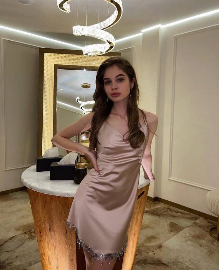 A woman in a pale pink satin slip dress with beaded fringe leans against a marble counter, with a gold-framed mirror and spiral chandelier visible in the background.
