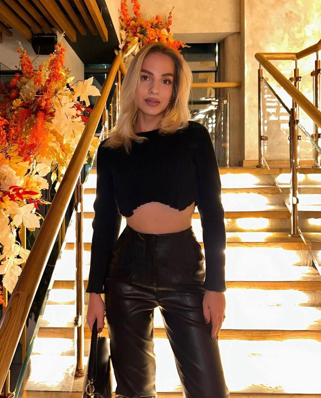 Zoya stands on a staircase in a black cropped knit top and leather pants, holding a handbag beside autumnal flowers.
