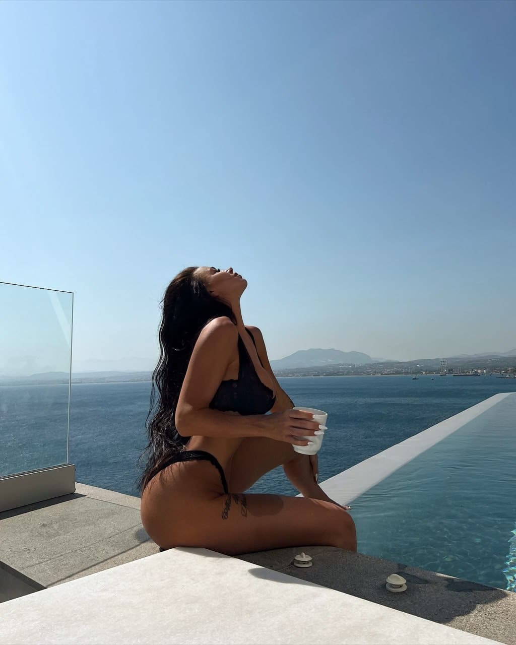 A woman sits on an infinity pool edge in a black bikini, holding a white cup and tilting her head back toward a blue sky over the sea.