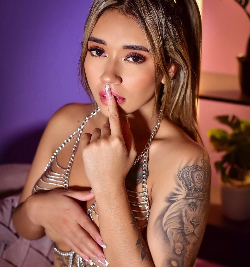 Woman in silver chain halter top touches her lips, posing in a room with purple and pink walls.