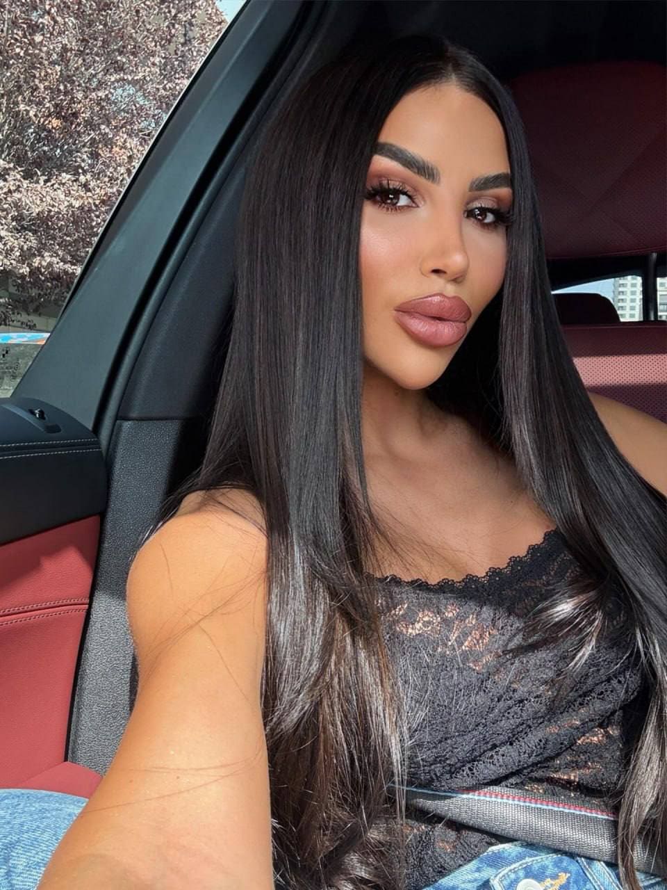 Sitting in a car's passenger seat wearing a black lace top, seatbelt across chest, red leather seats visible.