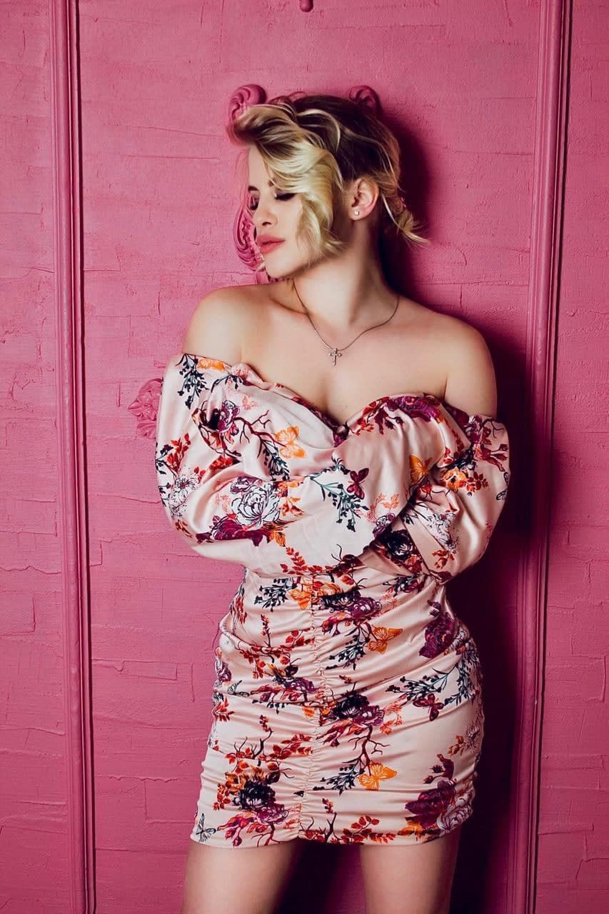 Standing with arms crossed against a pink wall, she wears a short off-the-shoulder floral dress.