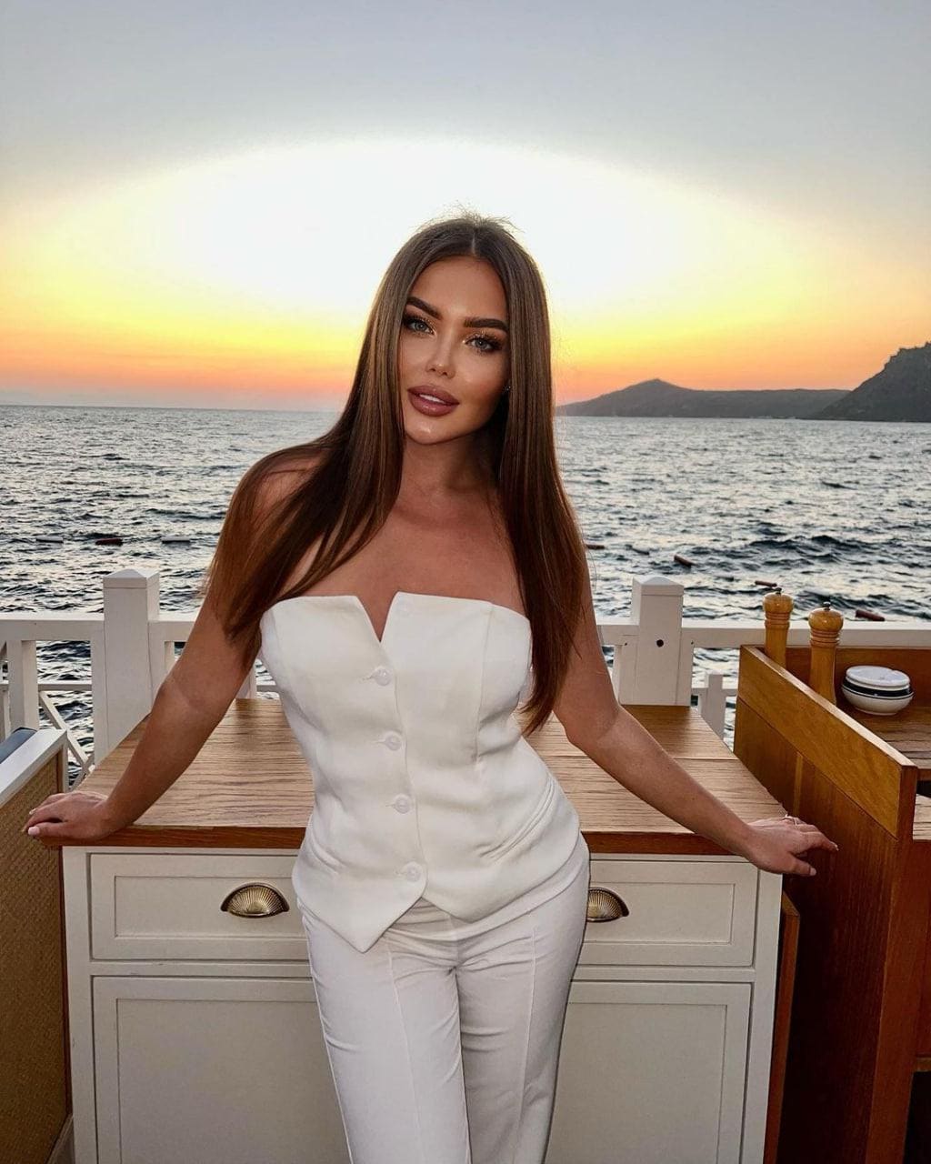 Женщина в белом топе без бретелей и брюках стоит на палубе лодки на закате, опираясь руками на деревянную стойку.