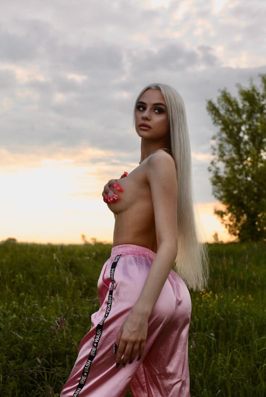 A woman in pink satin track pants stands in a grassy field at dusk, covering her chest with one hand and extending the other.