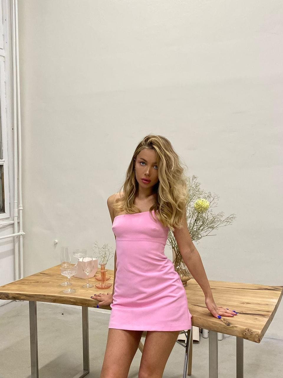 Zemfi leans on a wooden table with glassware, wearing a strapless pink mini dress in a white room.