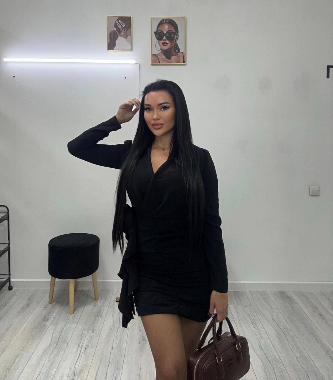 A woman in a black wrap dress holds a brown handbag in a room with light wood floors and framed portraits on the wall.