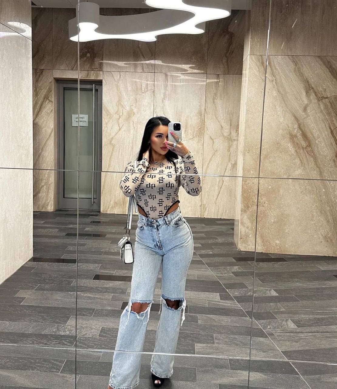 A mirror selfie in a marble lobby shows a woman in a patterned cropped top and ripped jeans, carrying a white shoulder bag.